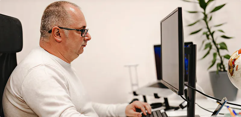 Man sitting at a desk with multiple computer monitors in an office setting.