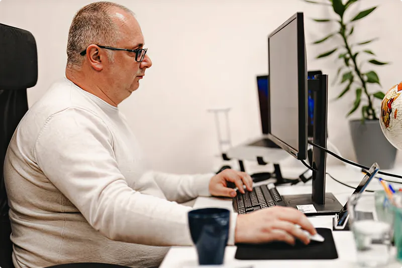 Man working at a desk with multiple computer monitors and office supplies.